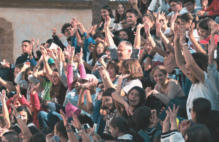 Un gruppo di persone partecipa al progetto "Fare comunit&agrave; insieme" della Fondazione con il Sud, nell'ambito del bando "Riabilitare il Sud". Le persone alzano le mani, creando un'atmosfera di partecipazione e coinvolgimento. L'evento si svolge all'aperto e coinvolge una folla di individui, alcuni dei quali indossano occhiali.