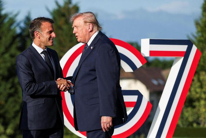 Donald Trump ed Emmanuel Macron sono riconoscibili nell'immagine mentre si stringono la mano. Entrambi indossano abiti formali, come giacche e completi. La scena si svolge all'aperto, davanti a un cartello.
