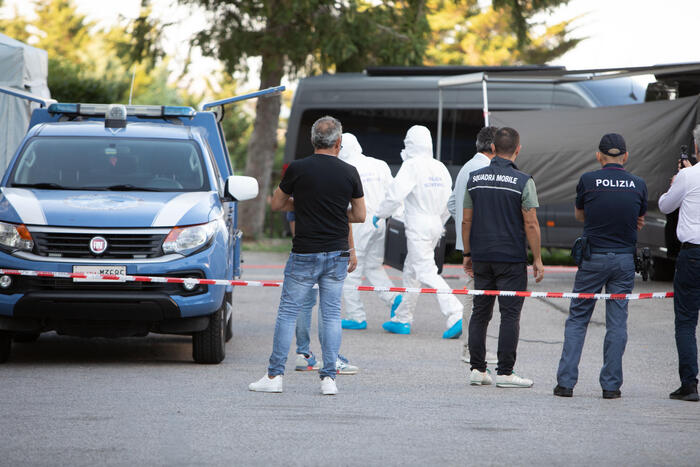 Un gruppo di persone in tute protettive &egrave; presente a Pietracatella, dove si &egrave; verificato un evento legato a una morte avvelenata. &Egrave; visibile anche un'auto blu con del nastro bianco e rosso. La scena &egrave; identificata come un luogo di crimine.