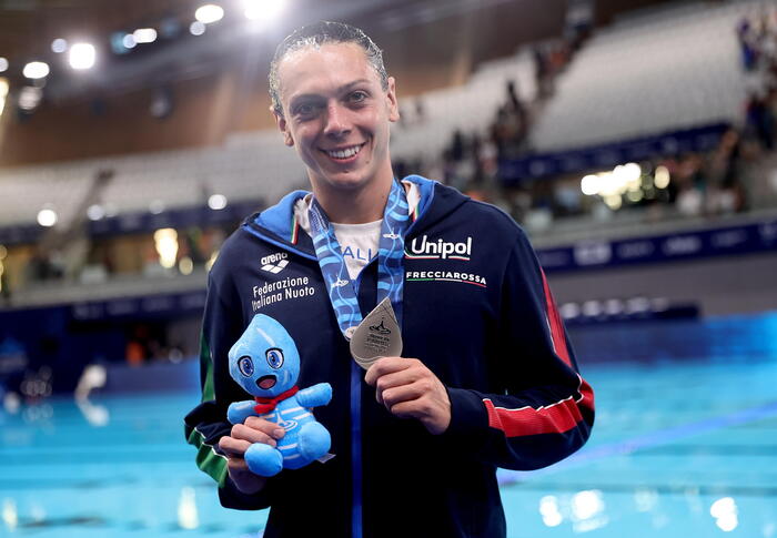 Un uomo partecipa ai Campionati Europei di Nuoto a Parigi nel 2026. Tiene in mano una medaglia e un piccolo animale di peluche blu. Indossa un abbigliamento sportivo, visibile insieme al suo volto.
