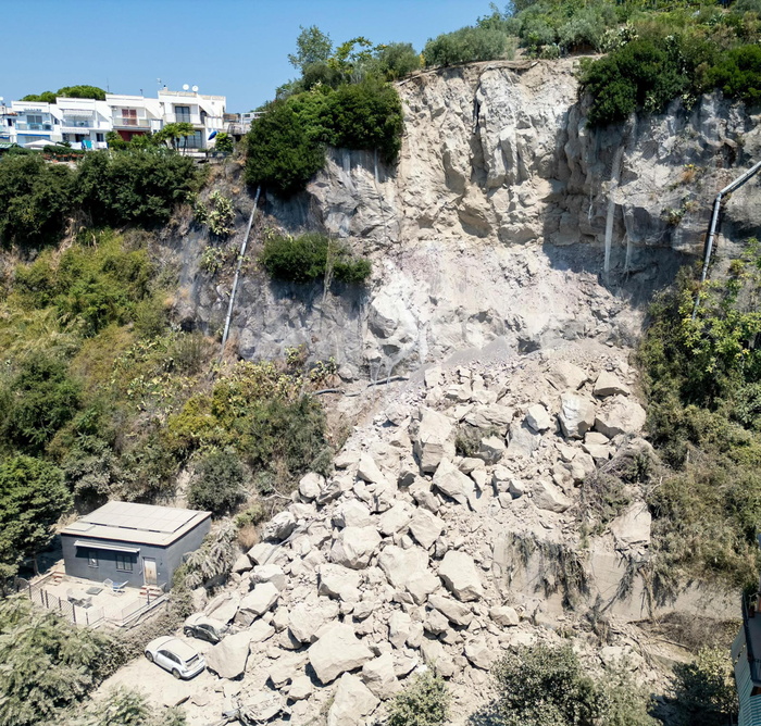 L'immagine rappresenta i Campi Flegrei, con accertamenti della Procura sulla messa in sicurezza dei costoni. Si osserva un costone roccioso con alberi e case. Il paesaggio include elementi naturali come piante e rocce, e presenta anche un edificio. Il contesto &egrave; all'aperto e caratterizzato da un ambiente naturale, con un rischio di frana evidenziato.