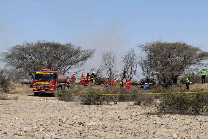 Un incidente aereo in Per&ugrave; ha causato la morte di 13 persone, tra cui 7 italiani. Nella scena, si osservano gruppi di persone in uniformi rosse e arancioni, probabilmente soccorritori o membri delle forze di emergenza. Sono presenti veicoli, tra cui un camion dei pompieri, in un contesto all'aperto con alberi e cielo visibile.