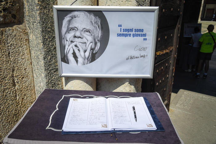 Si svolge il funerale di Don Mazzi nella Basilica di S. Ambrogio a Milano. Sono presenti un segno, una penna e un libro su un tavolo. Un testo significativo recita "sempre giovani" e "I sogni sono". La scena include anche una persona.