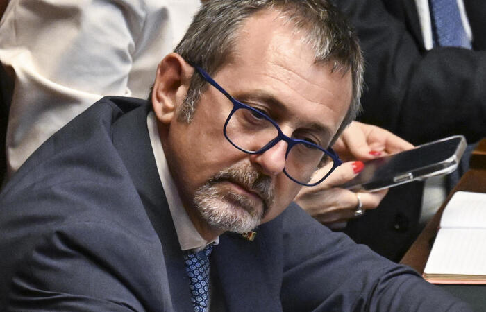 Andrea Delmastro &egrave; un uomo in giacca e cravatta seduto a una scrivania con un microfono. L'immagine si svolge in un ambiente interno.