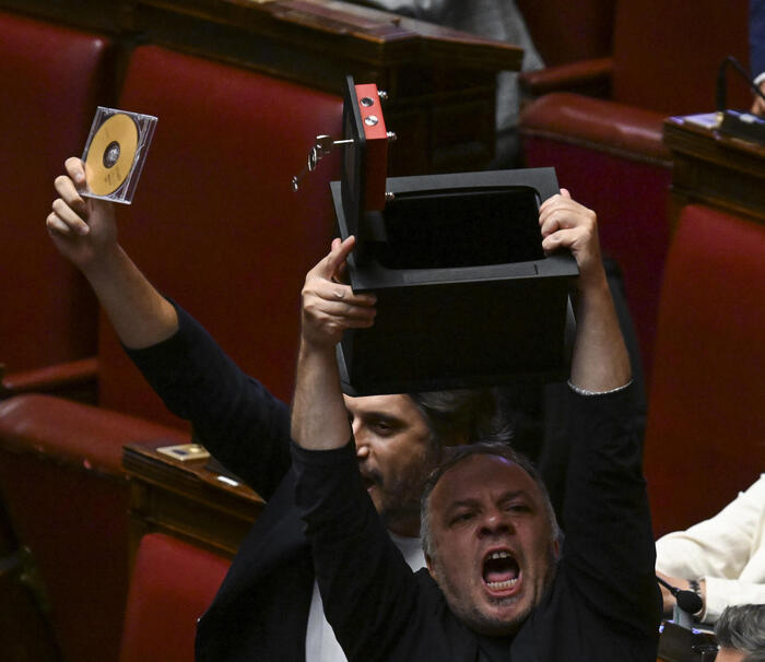 Un uomo tiene in mano una scatola e un CD. Il contesto mostra membri del M5S in Aula con striscioni che dicono "fuori le chat" e "aprite la cassaforte". Sono presenti altre persone, creando un'atmosfera di partecipazione. L'ambiente &egrave; interno e include elementi di arredamento.