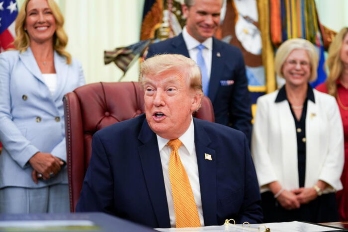 Donald Trump &egrave; riconoscibile nell'immagine. Annuncia un ordine esecutivo che stabilisce la Commissione per i Coniugi Militari Presidenziali. &Egrave; un uomo in abito formale, con giacca e cravatta. L'immagine presenta anche altre persone, suggerendo un contesto ufficiale e formale.