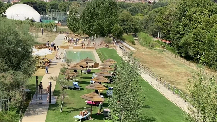 A Roma, nel Parco Tiberis, si osserva un'atmosfera estiva con un po' di sollievo per i bagnanti. L'immagine mostra una piscina circondata da un'area erbosa, con ombrelloni e persone che si godono il tempo all'aperto. Il parco &egrave; caratterizzato da alberi e una recinzione, creando un ambiente naturale e rilassante.