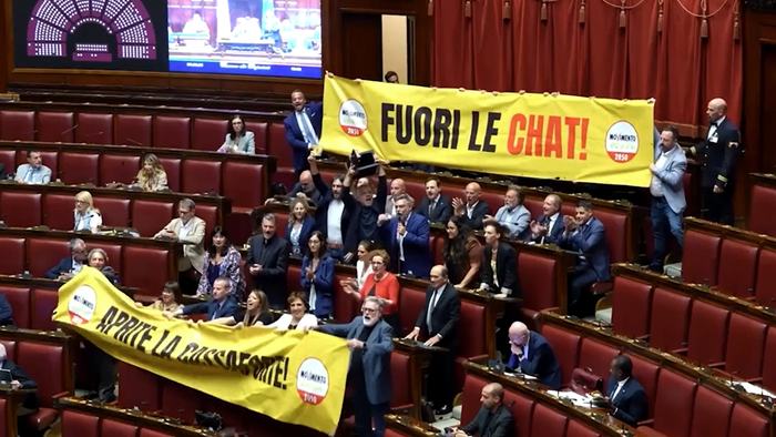 Un uomo tiene in mano una scatola e un CD. Il contesto mostra membri del M5S in Aula con striscioni che dicono "fuori le chat" e "aprite la cassaforte". Sono presenti altre persone, creando un'atmosfera di partecipazione. L'ambiente &egrave; interno e include elementi di arredamento.