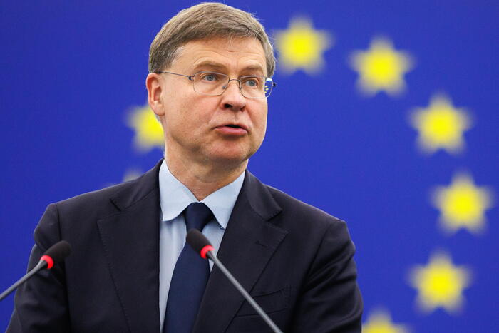 Valdis Dombrovskis &egrave; riconoscibile nell'immagine. &Egrave; un uomo in abito formale, con giacca e cravatta, che parla in un microfono durante una conferenza stampa. Dombrovskis discute temi come la presenza della Russia al G20 e la tassazione degli extraprofitti, in un contesto che coinvolge un pubblico e una folla di persone presenti.