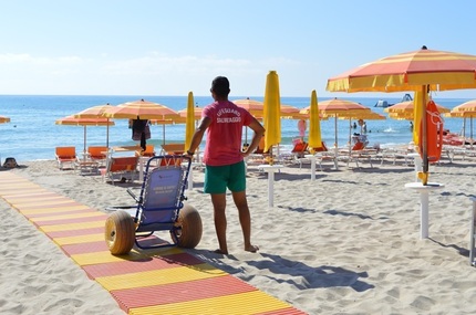 Un uomo spinge una sedia a rotelle su una spiaggia. La scena si svolge all'aperto, con il mare e la sabbia visibili. &Egrave; estate e il cielo &egrave; sereno. L'uomo &egrave; un adulto di sesso maschile. La spiaggia &egrave; un luogo di svago e natura, con acqua e terreno sabbioso.