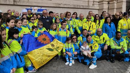 In Vaticano, un gruppo di persone in divise abbinati posa per una foto. Sono presenti due nuove ambulanze delle Misericordie, simbolo del volontariato. Il gruppo tiene anche una bandiera e indossa giacche luminose. La formazione &egrave; considerata il futuro del volontariato.