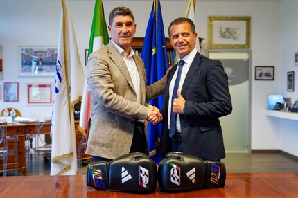 Due uomini si stringono la mano davanti a guantoni da boxe. L'immagine rappresenta un momento di accordo tra la Federazione Pugilistica Italiana (FPI) e il Comitato Italiano Paralimpico (CIP) per lo sviluppo della paraboxe. Sono presenti anche elementi come abbigliamento formale e un ambiente interno.