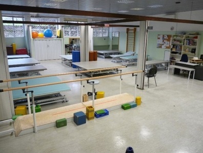 L'immagine rappresenta un ambiente per la riabilitazione nella sezione Aias di Monza. Si tratta di una stanza con una parete a specchio e tavoli. Sono presenti anche cubi sul pavimento. L'ambiente &egrave; all'interno di un edificio che funge da clinica e ospedale, con mobili e attrezzature tipiche di un laboratorio.