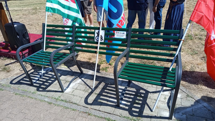 Nel Parco Carr&agrave; di Alessandria &egrave; inaugurata la Panchina Inclusiva. Un gruppo di persone si trova accanto a una panchina verde con gambe in metallo. La scena &egrave; all'aperto, su una superficie di mattoni, circondata da erba e piante.