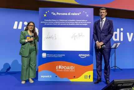 Amazon e il ministero per le Disabilit&agrave; firmano un protocollo d'intesa. Un uomo e una donna sono presenti accanto a un cartello blu con uno sfondo arancione. La scena include un pubblico formato da adulti, con persone in abbigliamento formale. Aggiuntivamente, &egrave; presente un testo significativo: "io, Persona di valore".