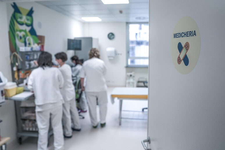 L'immagine mostra un reparto ospedaliero, con un gruppo di persone in una stanza. Tra di loro, si riconosce una persona che indossa pantaloni bianchi e un'altra figura sfocata con un camice bianco. L'ambiente &egrave; caratterizzato da elementi architettonici tipici di un ospedale, con attrezzature mediche e mobili presenti.