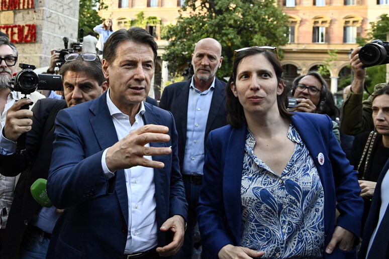 L'immagine rappresenta una maratona sui referendum, con i leader del Partito Democratico, del Movimento 5 Stelle, di Avs e di +E. Sono visibili un uomo e una donna, entrambi in abiti formali, con l'uomo che indossa un completo e la donna una giacca blu. L'ambientazione &egrave; all'aperto, e ci sono altre persone presenti. Un uomo tiene un telefono in mano.