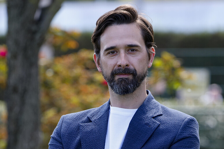 L'immagine mostra un uomo con barba e baffi, vestito con una giacca blu. Il suo volto &egrave; ben visibile, evidenziando i tratti distintivi. Il contesto &egrave; serio, poich&eacute; si fa riferimento a Davide Iacopini, che &egrave; in condizioni gravi, e a uno studente colpito. L'ambiente sembra essere all'aperto, con la presenza di alberi.