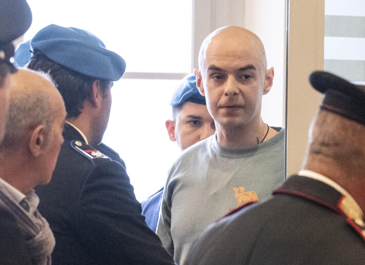 Il processo per l'omicidio del capotreno Alessandro Ambrosio ha inizio. Nell'immagine, &egrave; presente un uomo in camicia grigia circondato da uomini in uniforme. Altri uomini, tra cui uno in uniforme militare, sono visibili. L'ambiente suggerisce un contesto formale, probabilmente legato a questioni legali o ufficiali.