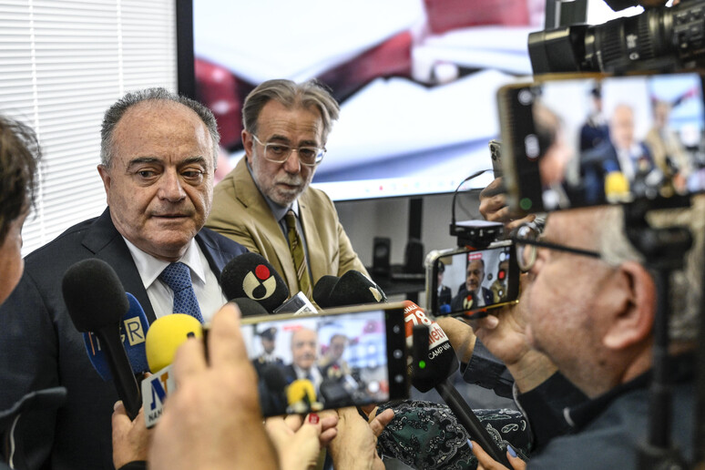 Durante una conferenza stampa, un gruppo di persone tiene in mano dei telefoni e parla con un uomo in giacca e cravatta. Un altro uomo, con un microfono, interagisce con i presenti. L'immagine mostra anche un operatore di telecamera e membri di una troupe televisiva. Il contesto riguarda la denuncia di Gratteri su dati rubati e venduti da poliziotti infedeli riguardanti calciatori e cantanti.