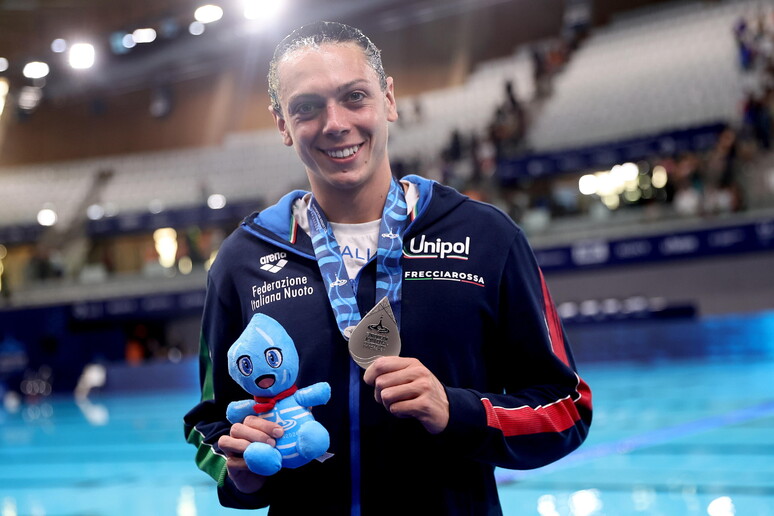 Un uomo partecipa ai Campionati Europei di Nuoto a Parigi nel 2026. Tiene in mano una medaglia e un piccolo animale di peluche blu. Indossa un abbigliamento sportivo, visibile insieme al suo volto.