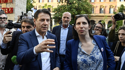 L'immagine rappresenta una maratona sui referendum, con i leader del Partito Democratico, del Movimento 5 Stelle, di Avs e di +E. Sono visibili un uomo e una donna, entrambi in abiti formali, con l'uomo che indossa un completo e la donna una giacca blu. L'ambientazione &egrave; all'aperto, e ci sono altre persone presenti. Un uomo tiene un telefono in mano.
