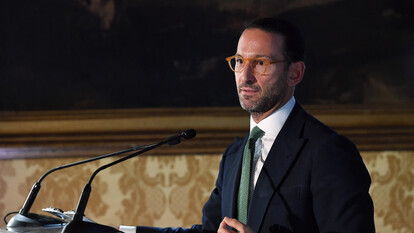 Un uomo in abito formale, con cravatta, si trova dietro a un podio. &Egrave; un oratore che sta parlando a un pubblico, composto da una folla di persone. Il contesto dell'immagine riguarda il rinvio della nomina del presidente della Consob.