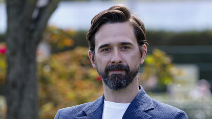 L'immagine mostra un uomo con barba e baffi, vestito con una giacca blu. Il suo volto &egrave; ben visibile, evidenziando i tratti distintivi. Il contesto &egrave; serio, poich&eacute; si fa riferimento a Davide Iacopini, che &egrave; in condizioni gravi, e a uno studente colpito. L'ambiente sembra essere all'aperto, con la presenza di alberi.
