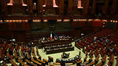 La scena si svolge in un'aula del Parlamento, caratterizzata da file di tavoli e sedie in legno. Si discute di una legge elettorale, con polemiche tra le opposizioni. Il Partito Democratico esprime preoccupazione per un possibile scontro istituzionale. Sono presenti anche 250 emendamenti in discussione. L'ambiente &egrave; interno e riflette un contesto di governo e architettura formale.