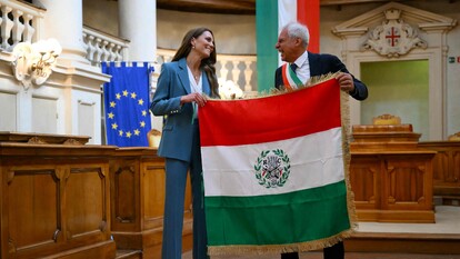 Kate &egrave; accolta in municipio a Reggio Emilia, dove riceve il primo Tricolore. Una donna e un uomo tengono in mano una bandiera, che presenta strisce verdi e bianche. Entrambi indossano abiti formali, inclusi un completo e un cappotto. L'evento si svolge all'interno di un edificio.