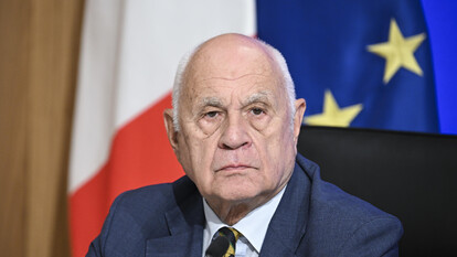 L'immagine ritrae un uomo in abito formale, con giacca e cravatta. &Egrave; un uomo adulto, identificato come un uomo d'affari. Il contesto suggerisce un ambiente ufficiale, probabilmente legato a un evento o a una situazione formale. La presenza di altre persone indica che si tratta di un contesto sociale o professionale.