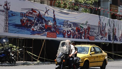 A Teheran, un uomo guida una motocicletta su una strada urbana. &Egrave; presente un banner con l'immagine di una nave. L'ambiente &egrave; caratterizzato da una strada con una recinzione e alberi. Sono visibili anche un'auto e altri veicoli. La scena riflette un contesto di conflitto tra Iran, Stati Uniti e Israele.