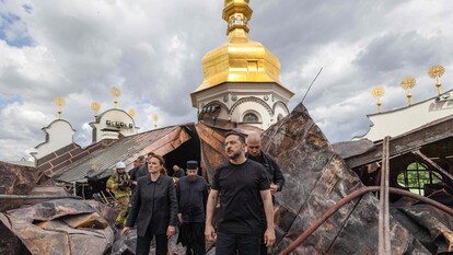 L'immagine mostra il presidente Zelensky sul luogo di un incendio, circondato da un gruppo di persone all'aperto. Si vede un edificio danneggiato, caratterizzato da un'architettura dorata e bianca con una torre. Il cielo &egrave; nuvoloso. Le persone indossano abbigliamento adatto per l'esterno.