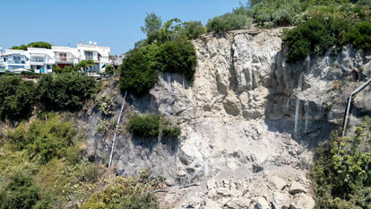 L'immagine rappresenta i Campi Flegrei, con accertamenti della Procura sulla messa in sicurezza dei costoni. Si osserva un costone roccioso con alberi e case. Il paesaggio include elementi naturali come piante e rocce, e presenta anche un edificio. Il contesto &egrave; all'aperto e caratterizzato da un ambiente naturale, con un rischio di frana evidenziato.