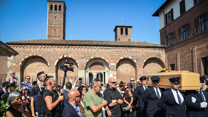 A Milano si svolgono i funerali di Don Mazzi, noto come il 'prete matto'. Un gruppo di persone &egrave; presente all'esterno di un edificio. Tra di loro ci sono uomini in abiti formali, alcuni in giacca e cravatta. L'atmosfera &egrave; caratterizzata dalla presenza di paparazzi e da un cielo visibile sopra la scena.