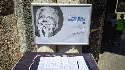 Si svolge il funerale di Don Mazzi nella Basilica di S. Ambrogio a Milano. Sono presenti un segno, una penna e un libro su un tavolo. Un testo significativo recita "sempre giovani" e "I sogni sono". La scena include anche una persona.