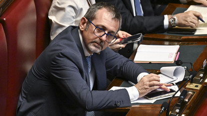 Andrea Delmastro &egrave; un uomo in giacca e cravatta seduto a una scrivania con un microfono. L'immagine si svolge in un ambiente interno.