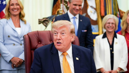 Donald Trump &egrave; riconoscibile nell'immagine. Annuncia un ordine esecutivo che stabilisce la Commissione per i Coniugi Militari Presidenziali. &Egrave; un uomo in abito formale, con giacca e cravatta. L'immagine presenta anche altre persone, suggerendo un contesto ufficiale e formale.