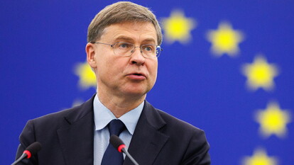 Valdis Dombrovskis &egrave; riconoscibile nell'immagine. &Egrave; un uomo in abito formale, con giacca e cravatta, che parla in un microfono durante una conferenza stampa. Dombrovskis discute temi come la presenza della Russia al G20 e la tassazione degli extraprofitti, in un contesto che coinvolge un pubblico e una folla di persone presenti.