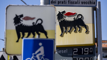 L'immagine mostra un distributore di carburante Eni, dove il prezzo del gasolio &egrave; stato ridotto a 2,19 euro. Sono visibili segnali stradali, inclusi cartelli con un cane e indicazioni di prezzo. L'ambiente &egrave; all'aperto, con un cielo visibile.